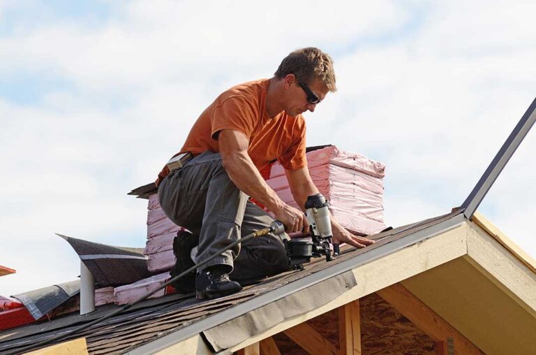 A person placing a bucket under a dripping ceiling next to furniture covered with a tarp.